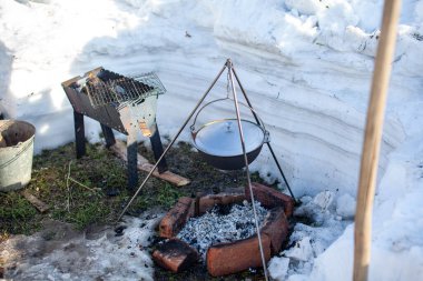 Ateşin üzerinde yemek pişirmek için bir tencere asılı. Üç ayaklı bir kancada, tavadan buhar çıkıyor. Kış kampı açık hava yemekleri.