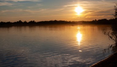 Sakin bir gölde güneşli, renkli bir gün batımı. Güneş suyun yüzeyine yansıyor..