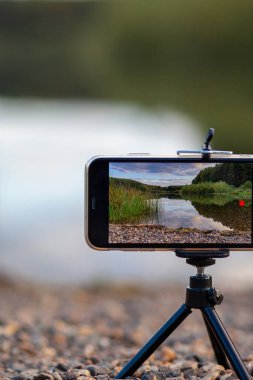 Bir tripodun üzerindeki telefonun yakın çekimi doğanın bir videosunu ya da fotoğrafını çeker. Ormanda güzel bir göl. Fotoğrafçının cep telefonunda bulutlar var.. 