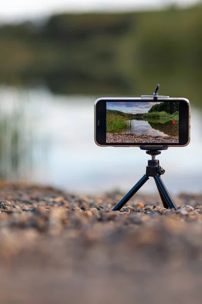 Bir tripodun üzerindeki telefonun yakın çekimi doğanın bir videosunu ya da fotoğrafını çeker. Ormanda güzel bir göl. Fotoğrafçının cep telefonunda bulutlar var.. 
