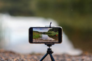 Bir tripodun üzerindeki telefonun yakın çekimi doğanın bir videosunu ya da fotoğrafını çeker. Ormanda güzel bir göl. Fotoğrafçının cep telefonunda bulutlar var.. 