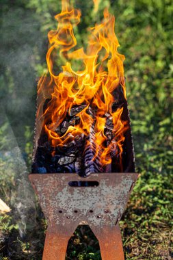 Izgaradaki yakacak odun, doğal yeşil arka planda parlak turuncu bir ateş alevi ile yanıyor. Doğada ızgarada et pişirme hazırlığı. Ateş ve duman 