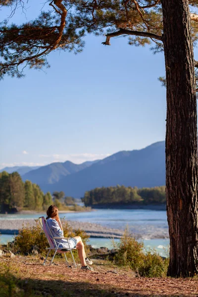 A woman is sitting on a folding chair on the bank of a mountain river on a nice, warm day under a large tree. A calm and quiet place to relax and reflect. Equipment and a tourist\'s rest.