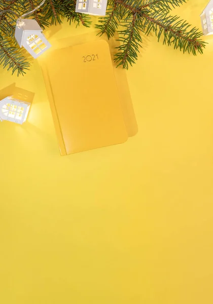 Świąteczne miejsce pracy świąteczne tło pionowe: notatnik na 2021 r., gałązki jodły, błyszczące lampki świąteczne białe domki na żółtym tle. — Zdjęcie stockowe