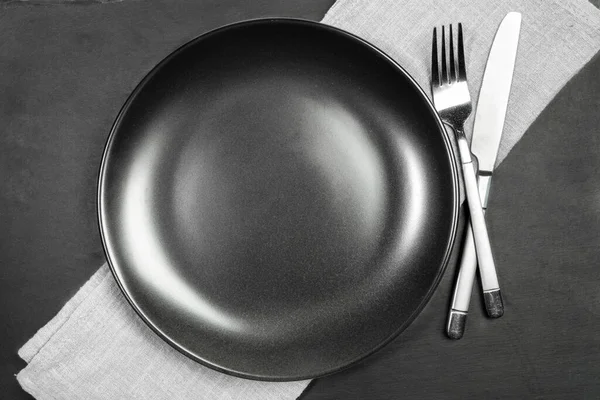 Assiette noire vide et couverts sur une serviette de cuisson en diagonale grise sur une planche de service en schiste noir. — Photo