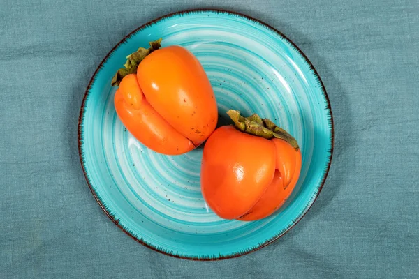 Two ugly orange persimmons on turquoise plate on dark turquoise textile backdrop. Zero waste food. Fresh healthy organic fruits. Reasonable sustainable consumption. Top view, close up.