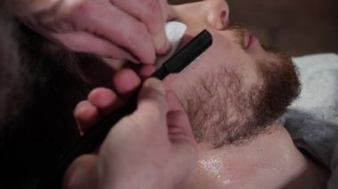 Genç müşterinin berber dükkanında sakalını jiletle tıraş edişinin yakın çekimi.