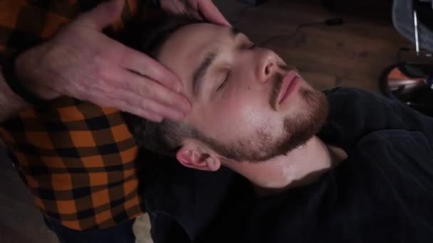 Парикмахер массирует лицо молодого клиента после стрижки — стоковое видео
