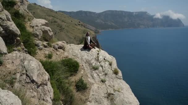 Uzun saçlı genç bir adam uçurumun kenarında meditasyon yapıyor. — Stok video