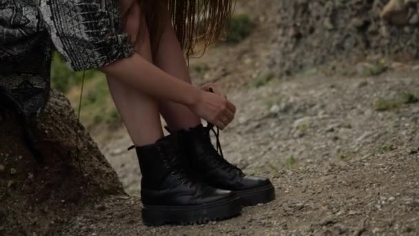 Una chica con una capa gris se sienta en una piedra y se ata los cordones de los zapatos — Vídeos de Stock