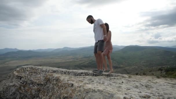 Pareja joven enamorada se eleva al borde de un acantilado contra las montañas de telón de fondo — Vídeo de stock