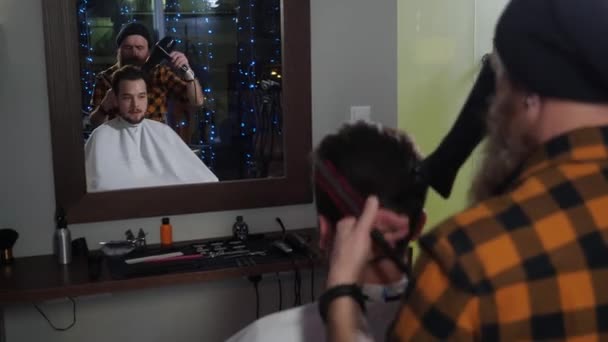 Volwassen baardkapper maakt styling een jonge klant haardroger in een kapperszaak — Stockvideo