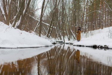 Uzun ceketli genç bir kız kış ormanında nehrin kenarında duruyor.
