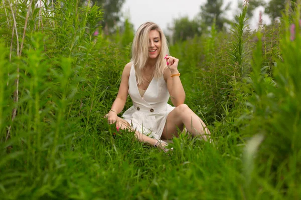 Яскрава блондинка в білій сукні і з червоним манікюром сидить на траві — стокове фото
