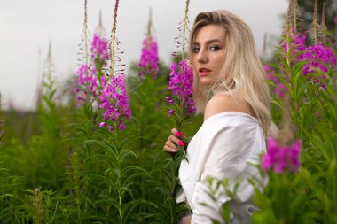 Beyaz gömlekli parlak sarışın kız çiçek açan çayırların arasında duruyor.