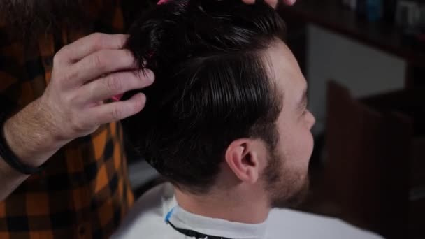 Мужчина парикмахер расчесывает волосы розовые волосы для парня клиента, который сидит в кресле — стоковое видео