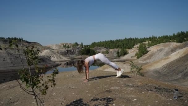 En ung flicka i vit sportuniform gör lungor och hoppar på ett sandberg — Stockvideo