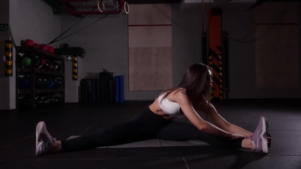 Стройная девушка в черных леггинсах и фиолетовых кроссовках делает растяжку в спортзале — стоковое видео