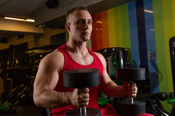 Fiziği olan yetişkin bir adam spor salonunun simülatörüne oturmuş, elinde dambıllar tutarak ileriye bakıyor. Yüksek kalite fotoğraf