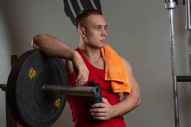 Kırmızı tişörtlü bir erkek sporcu spor salonunda duruyor ve simülatöre yaslanıyor. 