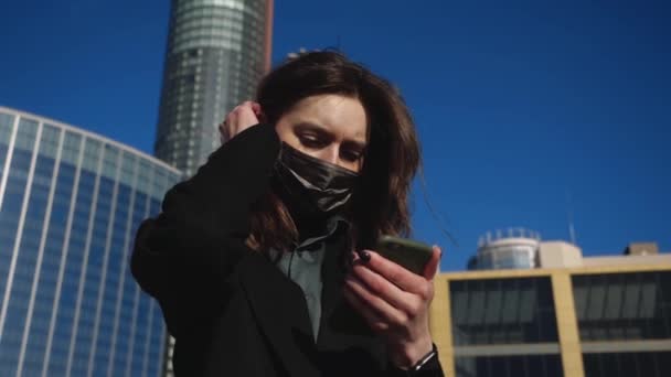 Κορίτσι σε μια προστατευτική μάσκα κρατά ένα κινητό τηλέφωνο στο παρασκήνιο της μεγάλης πόλης — Αρχείο Βίντεο