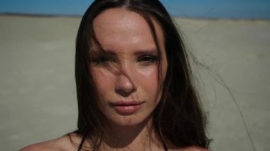 Güzel bir kız kumlu bir sahilde duruyor ve seksi bir şekilde kameraya bakıyor.