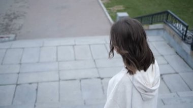 Kahverengi saçlı genç bir kız merdivenlerden inerek şehirde dolaşıyor.