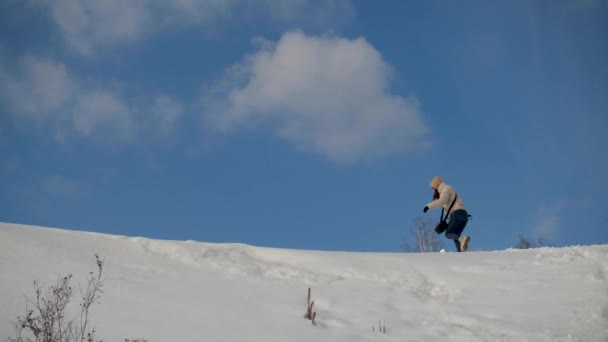 Ένα κορίτσι ταξιδιώτισσα με σακάκι σκαρφαλώνει σε ένα χιονισμένο βουνό στον συννεφιασμένο ουρανό — Αρχείο Βίντεο