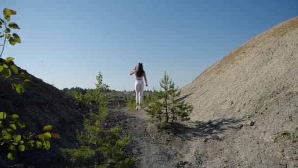 A girl in a sporty uniform walks along a sandy mountain road towards the lake — 图库视频影像