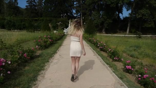 En förförisk flicka med långt blont hår går längs en blomstrande gränd — Stockvideo