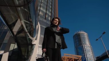 Elinde deri evrak çantası olan bir kız yüksek bir ofis binasının yanında duruyor.