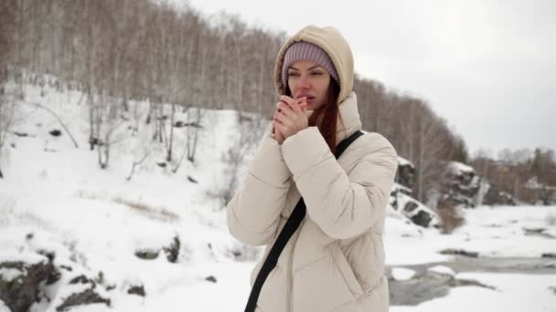 A beautiful girl in a warm jacket and a hood is trying to warm frozen hands — Stock video