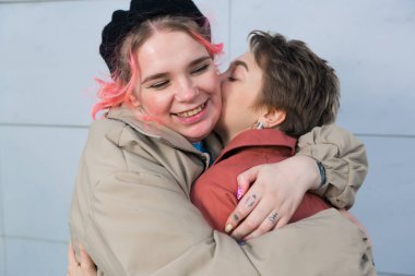 Bir çift genç kız birbirlerine sarılır ve ayakta güler. 