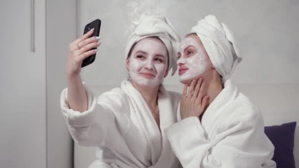 Две девушки в халатах и с глиняными масками на лицах делают селфи — стоковое видео