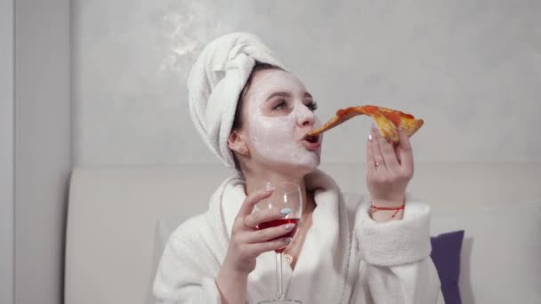 Dziewczyna w ręczniku na głowie i w białym szlafroku pije czerwone wino i je pizzę — Wideo stockowe