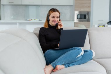 Genç ve güzel bir kız laptopun arkasında çalışıyor kulaklıkla konuşuyor, evde oturuyor.