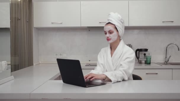 Uma menina em um manto branco e máscara facial funciona atrás de um laptop sentado em casa — Vídeo de Stock