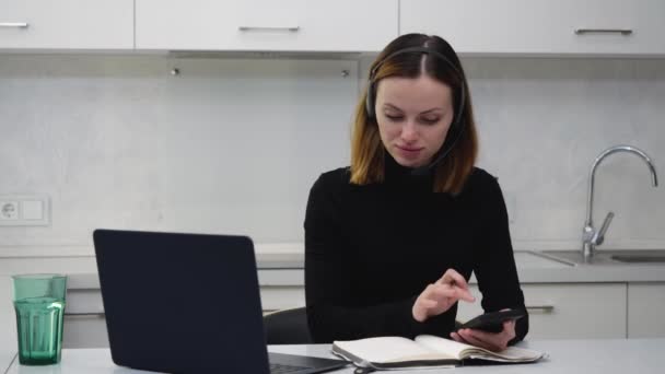 Una joven en un auricular toma notas en su teléfono y portátil — Vídeo de stock