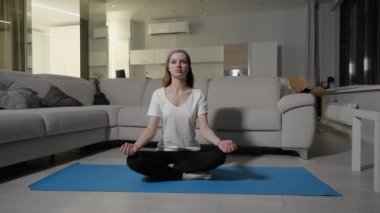 Beyaz tişörtlü genç bir kız evdeki mavi yoga minderinde lotus pozunda oturuyor.