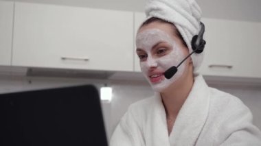 Bornozlu bir kız kulaklıkla neşeli bir şekilde konuşuyor, evde oturuyor.