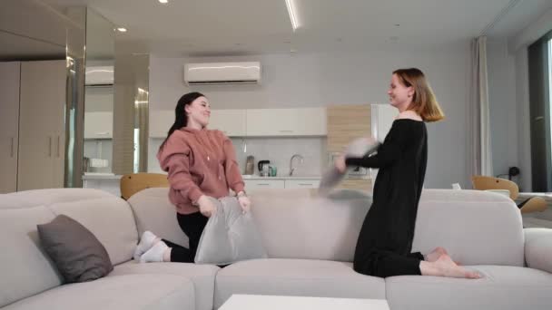 Young girls cheerfully fight pillows at home on the couch — Stock Video