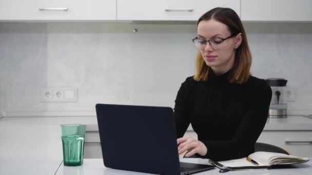Chica inteligente con gafas trabaja detrás de un ordenador portátil y toma notas en un cuaderno — Vídeo de stock