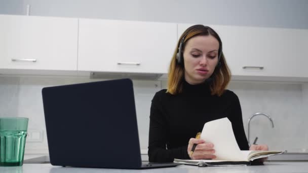 Молода розумна дівчина розмовляє по гарнітурі, роблячи нотатки в блокноті — стокове відео