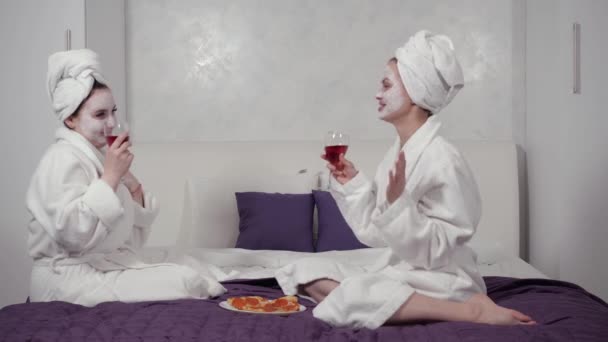 Dois amigos bebem vinho e enlouquecem com óculos sentados em roupões na cama — Vídeo de Stock