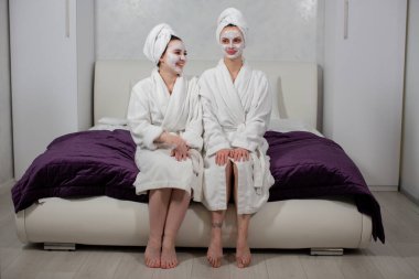 Kafalarında havlular, yüzlerinde maske olan kadınlar yatağa otururlar.