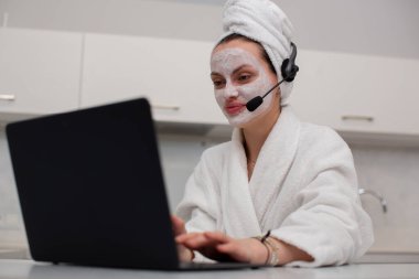 Sabahlık giymiş bir kız, kulaklıkla dizüstü bilgisayarın arkasında çalışıyor.