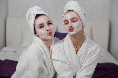 Nemlendirici yüz maskeli, beyaz cüppeli iki kız yatakta oturuyor.