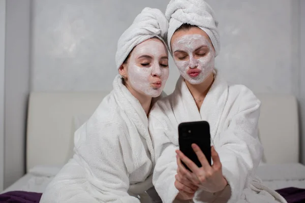 Két vicces lány barátnő fürdőköpenyben, arcmaszkok szelfik a telefonban. — Stock Fotó