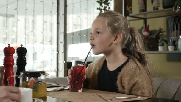 A little girl with blonde hair sitting in a restaurant drinking juice — Stock Video