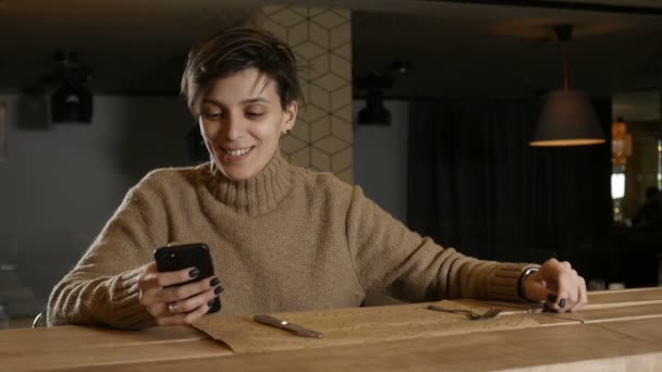 Osamělá dívka sedí v restauraci, usmívá se a dívá se na telefon — Stock video